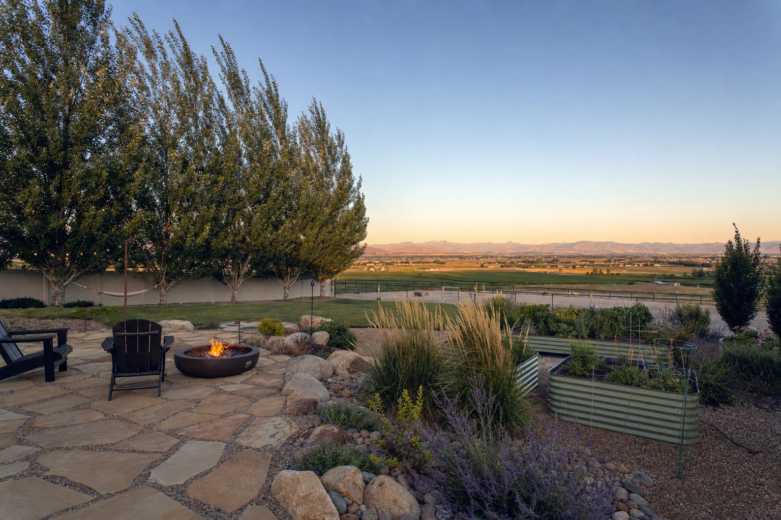 Kuna, Idaho rural-suburban backyard landscape with flagstone patio, fire pit, raised vegetable garden beds, mature Siberian elm windbreak trees, drought-tolerant ornamental grasses and Russian sage, with distant views of agricultural fields and the Owyhee Mountains under a high-desert summer evening sky